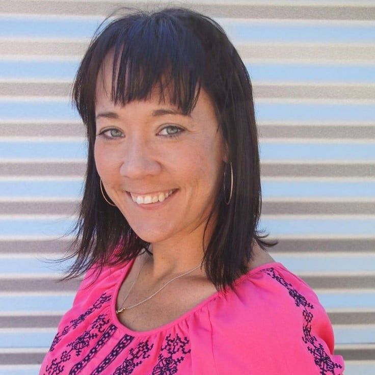 Bio photo of Kristen Auerbach, smiling in a pink shirt against a corrugated metal background